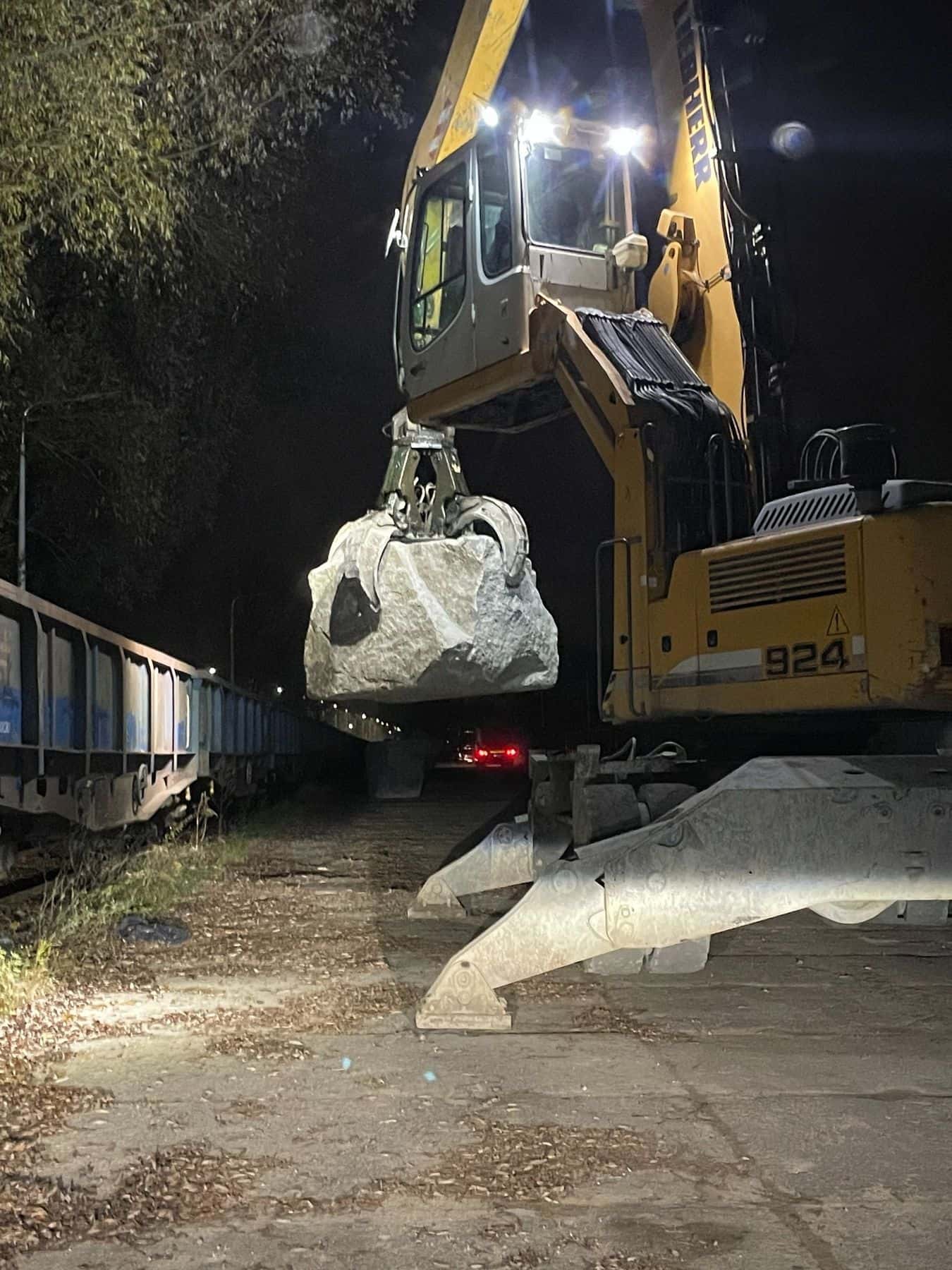 Koparka przeładunkowa podnosi bryłę kamienia hydrotechnicznego nad wagonem podczas nocnego załadunku