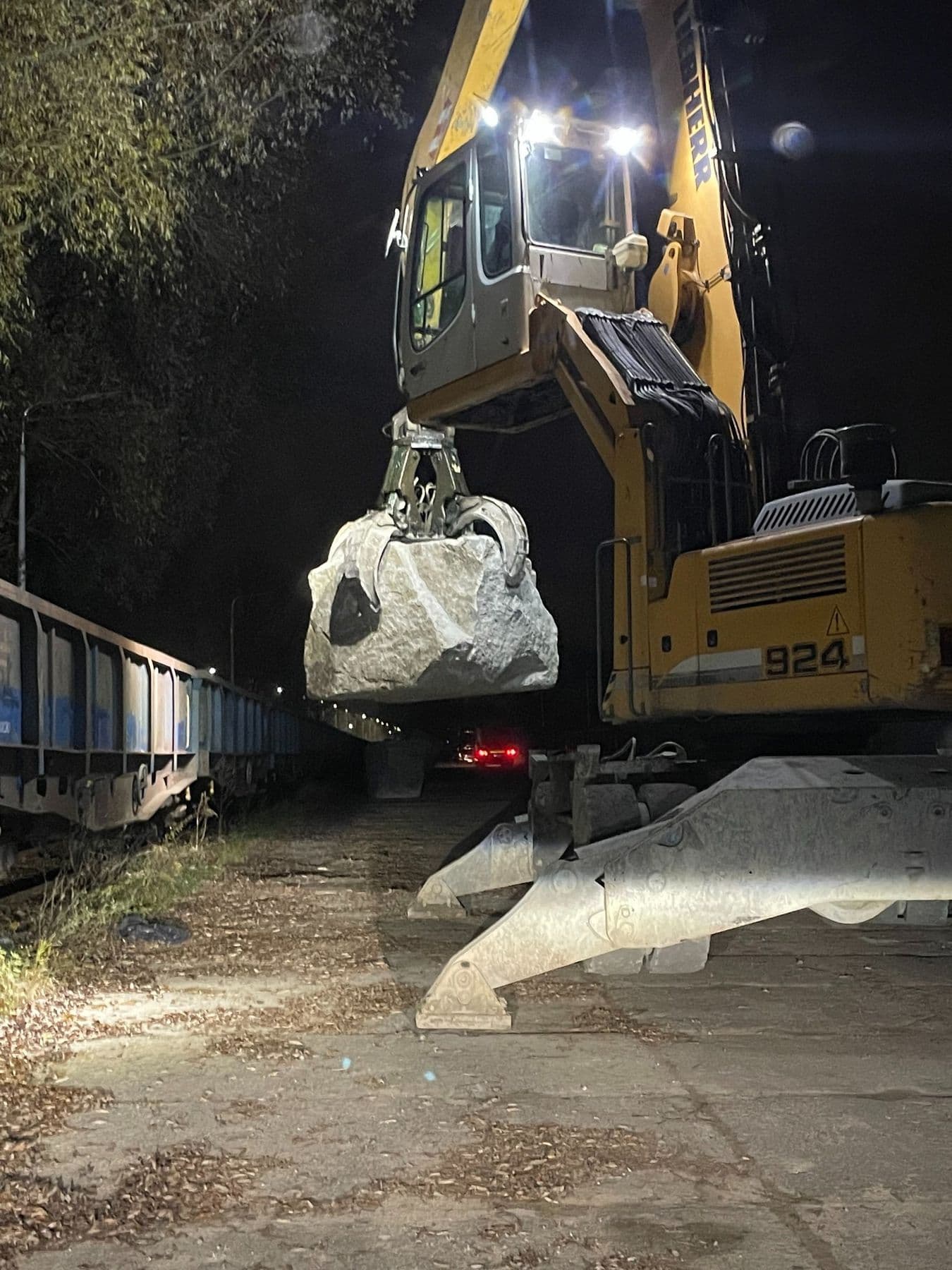 Koparka przeładunkowa podnosi bryłę kamienia hydrotechnicznego nad wagonem podczas nocnego załadunku